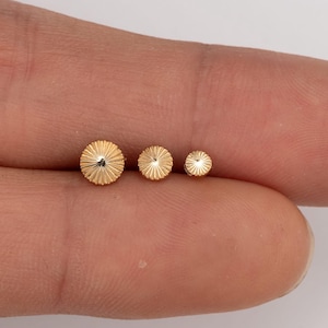 May include: Three small, gold-colored, round earrings with a sunburst design are displayed. The earrings are of varying sizes, with the largest measuring approximately 0.25 inches in diameter. The earrings are set against a neutral background.