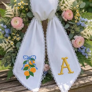 Peut inclure: Écharpe de baptême en lin blanc avec broderie d'oranges et de feuilles, un nœud bleu et la lettre "A" en or. L'écharpe est drapée sur une couronne florale de roses roses, de fleurs bleues et de verdure, posée sur un banc en bois.