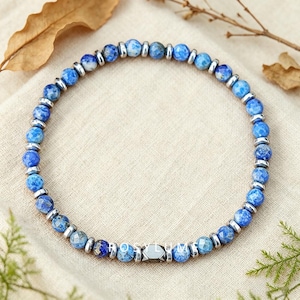 Pode incluir: Uma pulseira de contas com alternância de contas de pedra azul redondas e facetadas e pequenos espaçadores metálicos lisos de tom prateado. A pulseira está disposta em círculo e inclui uma conta geométrica central de metal escuro.