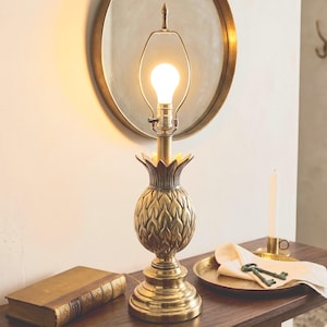 Puede incluir: Una lámpara de mesa de latón con forma de piña con una bombilla encendida. La lámpara está sobre una mesa de madera, junto a un libro, una vela y una bandeja decorativa con llaves. Un espejo redondo cuelga en la pared del fondo.