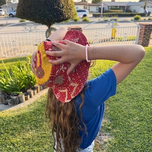 May include: A red and yellow patterned hat with a floral design. The hat is held up by a person wearing a blue t-shirt and light blue jeans. A pink and white beaded bracelet is visible on the person's wrist.