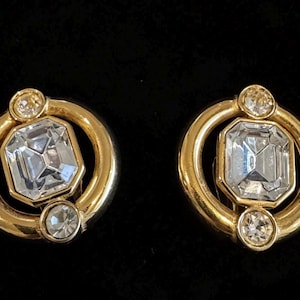 Puede incluir: Un par de pendientes circulares de tono dorado con un cristal transparente central de corte esmeralda rodeado por un anillo de metal liso. Cada pendiente está decorado con dos cristales redondos transparentes más pequeños arriba y abajo.