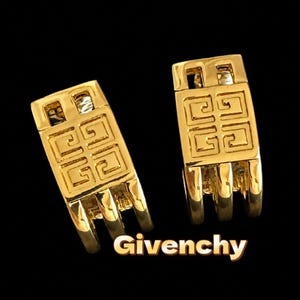 May include: A pair of gold-toned metal earrings featuring a rectangular design with an embossed geometric logo pattern and three vertical prongs at the base. The word Givenchy is printed in white text at the bottom of the image.