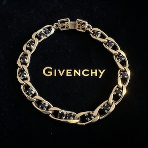 Puede incluir: Una pulsera de eslabones dorados con piedras negras de corte cuadrado alternadas en los eslabones. La pulsera incluye un cierre rectangular y está colocada sobre el texto dorado GIVENCHY impreso en un fondo oscuro.
