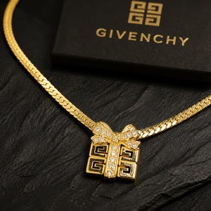 Puede incluir: Un collar de cadena de espiga en tono dorado con un colgante en forma de caja de regalo con lazo. El colgante está adornado con cristales transparentes y muestra el logotipo de Givenchy. El collar se sitúa junto a una caja negra Givenchy.