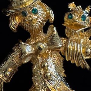 Könnte beinhalten: Eine goldfarbene Metallbrosche, die eine Vogelscheuche mit Hut und Fliege darstellt, mit zwei kleinen grünen Strasssteinen als Augen. Ein kleiner Vogel mit zwei grünen Strassaugen sitzt auf der Schulter der Vogelscheuche. Die strukturierte Metalloberfläche hat ein helles, reflektierendes Finish.
