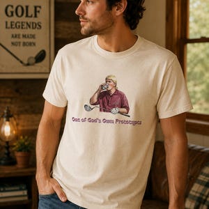 May include: Beige t-shirt featuring a graphic of a golfer drinking from a glass, with the text "One of God's Own Prototypes" in purple. The shirt is worn by a person in blue jeans.