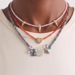 May include: Three layered necklaces with different designs. The top necklace is pink with gold spikes. The middle necklace is red with a gold heart pendant. The bottom necklace has a flower and leaf pendant with colorful beads.