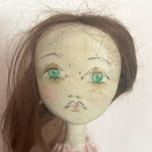 May include: A doll head with a pale, off-white painted surface featuring hand-drawn facial details including bright green eyes, thin eyebrows, and pink cheeks. The doll has long, thin, brown hair strands scattered across the top and sides.