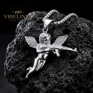 Bold Cupid angel necklace with cubic zirconia crystal wings in gold and silver stainless steel, laying side by side on a dark background, a striking streetwear statement piece and unique gift for jewelry collectors.