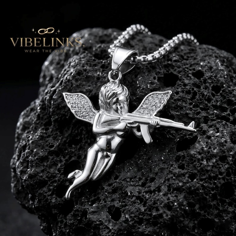 Bold Cupid angel necklace with cubic zirconia crystal wings in gold and silver stainless steel, laying side by side on a dark background, a striking streetwear statement piece and unique gift for jewelry collectors.