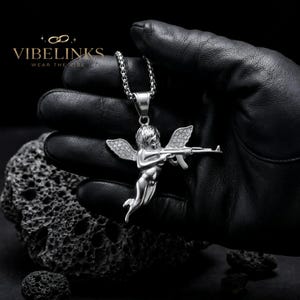 Silver tone Cupid angel pendant necklace in stainless steel with iced-out crystal wings, photographed flat on a dark surface alongside its gold counterpart, a luxury-looking hip hop jewelry piece with 60cm chain included.