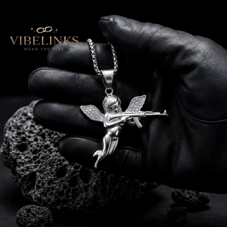 Silver tone Cupid angel pendant necklace in stainless steel with iced-out crystal wings, photographed flat on a dark surface alongside its gold counterpart, a luxury-looking hip hop jewelry piece with 60cm chain included.