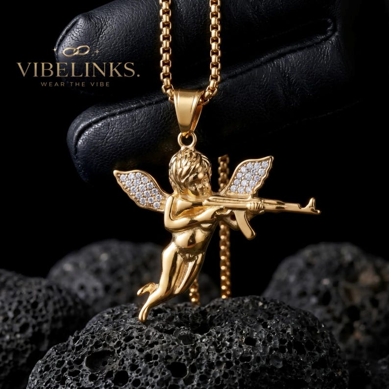 Side-by-side gold and silver Cupid angel necklaces with crystal wings on a dark fabric surface, showing both color variations of the stainless steel CZ angel charm, a bold unisex jewelry gift for men and women.
