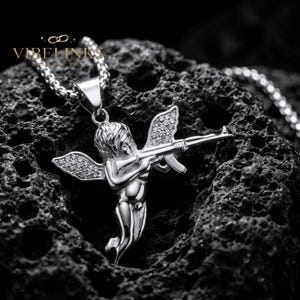 Unisex Cupid angel pendant necklace in gold and silver tone stainless steel with CZ crystal wings, photographed on a dark surface — a versatile statement jewelry piece for layering, everyday wear, or as a bold fashion gift.