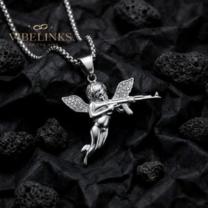Two stainless steel Cupid angel pendant necklaces displayed on a dark textured surface, one in gold tone and one in silver tone, both featuring a flying Cupid figure with pavé cubic zirconia crystal wings, hanging from 60cm box chains.