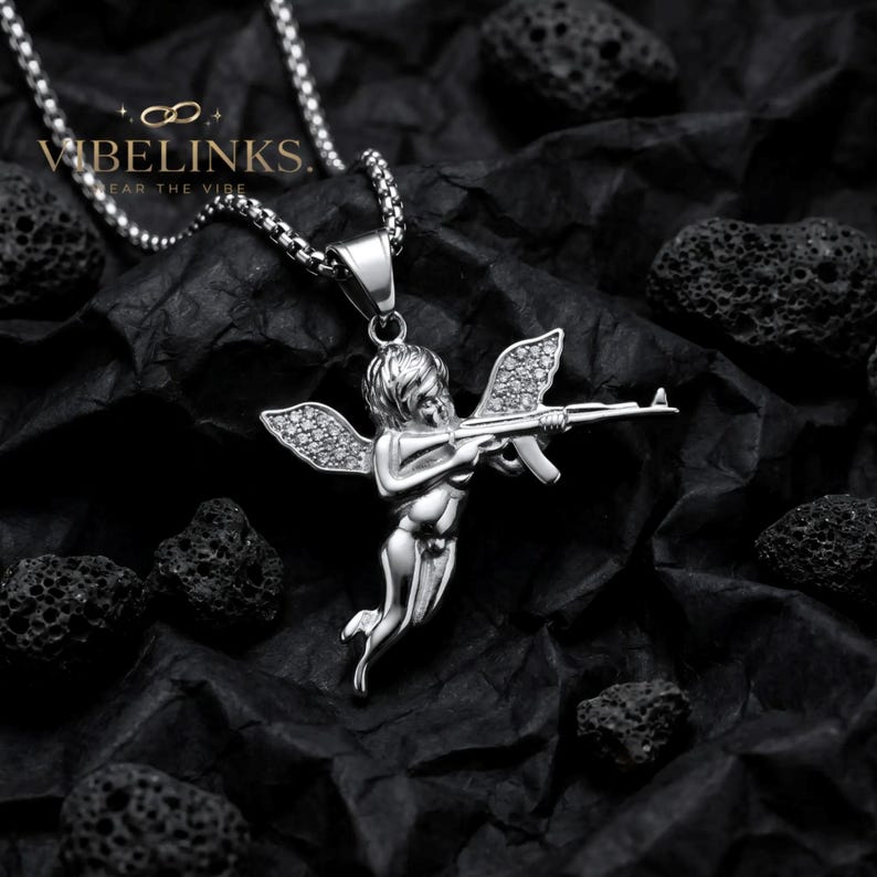 Two stainless steel Cupid angel pendant necklaces displayed on a dark textured surface, one in gold tone and one in silver tone, both featuring a flying Cupid figure with pavé cubic zirconia crystal wings, hanging from 60cm box chains.