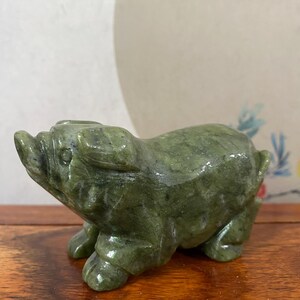 Puede incluir: Una pequeña figura de cerdo tallada a mano en una pieza sólida de piedra verde oliva moteada. La escultura presenta una superficie lisa y pulida con variaciones minerales naturales visibles, representando al animal en posición de pie.
