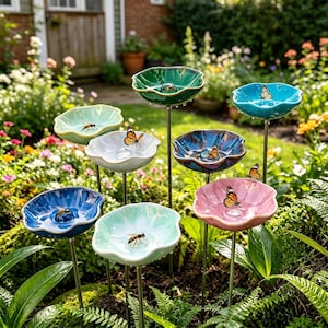 Könnte beinhalten: Eine Sammlung farbenfroher Vogeltränken aus Keramik in einem Garten. Jede Tränke ist wie eine Blume geformt und auf einem Metallstab montiert. Schmetterlinge und Bienen sitzen auf den wassergefüllten Schalen. Die Tränken sind in Blau-, Grün- und Rosatönen gehalten.