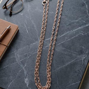 Op de afbeelding: Roségouden ketting met karabijnsluitingen, bril met schildpadmontuur en een bruin leren notitieboek met pen op een donkergrijze marmeren ondergrond.