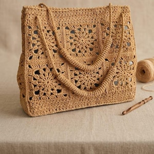 May include: A tan crochet tote bag constructed from multiple square motifs featuring a central floral pattern. The bag includes two matching shoulder straps and an open top design. A wooden crochet hook and a small ball of yarn are placed nearby.