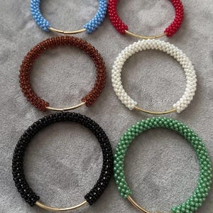 May include: Six circular bracelets arranged in two columns, each featuring a gold-toned metal wire segment and a continuous loop of small, uniform beads in light blue, red, dark brown, white, black, and green, creating a textured appearance.