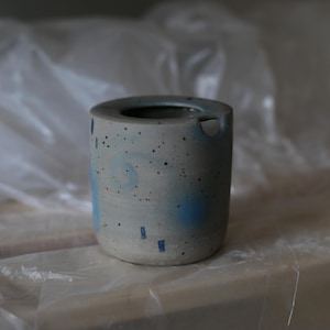 Puede incluir: Un vaso cilíndrico de cerámica con base gris claro, que presenta motas oscuras, acentos circulares azules y pequeñas marcas rectangulares azules. El vaso tiene un borde redondeado y liso y una pequeña abertura circular cerca del borde.