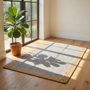 Peut inclure: Un tapis rectangulaire composé d'un panneau central tissé gris clair encadré par une bordure épaisse et texturée de couleur beige naturel. Le tapis est fabriqué à partir de fibres tressées et présente l'ombre portée d'une plante.