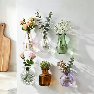 Peut inclure: Une collection de vases en verre muraux de différentes formes et couleurs, chacun contenant différents types de fleurs et de verdure. Les vases comprennent des teintes roses, claires, vertes et violettes, créant un affichage floral décoratif. L'ensemble est posé sur un mur blanc.