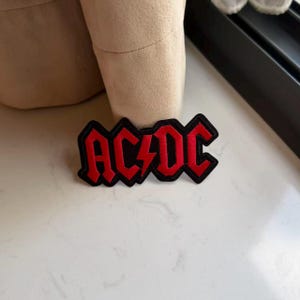 Può includere: Una toppa ricamata rettangolare che presenta il testo AC/DC in grassetto rosso con un simbolo centrale a forma di fulmine. Le lettere sono delineate con filo nero, creando un netto contrasto con il riempimento rosso vivace del logo.