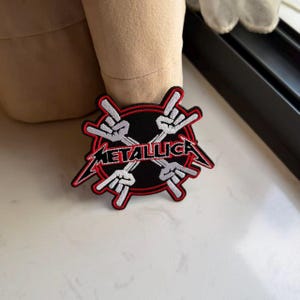 May include: A circular embroidered patch featuring the Metallica logo in black and white with red outlines. The design includes four white hands in a rock-and-roll gesture arranged in a cross pattern over a black circular background with a red border.