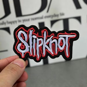 May include: Embroidered patch featuring the band name "Slipknot" in white, outlined in red, on a black background. The patch has a unique, stylized font and is likely designed for application on clothing or accessories.