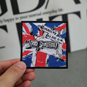 May include: Embroidered patch featuring the Union Jack flag design with the words "Sex Pistols" and "Anarchy in the U.K." in a distressed style. The patch has a black border and is square-shaped.