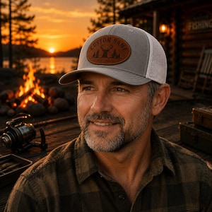 Puede incluir: Gorra trucker gris con parte trasera de malla blanca y parche de cuero marrón. El parche presenta una silueta de ciervo, árboles y el texto "CUSTOM NAME" y "EST. 2024". La gorra la lleva una persona.