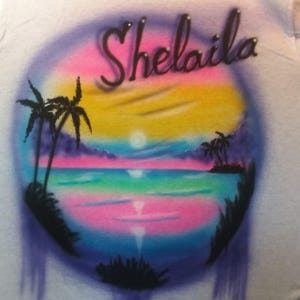 May include: White t-shirt with an airbrushed tropical sunset design. The design features a colorful sky with pink, yellow, and blue hues, palm trees, and the name "Shelaila" in a cursive font.