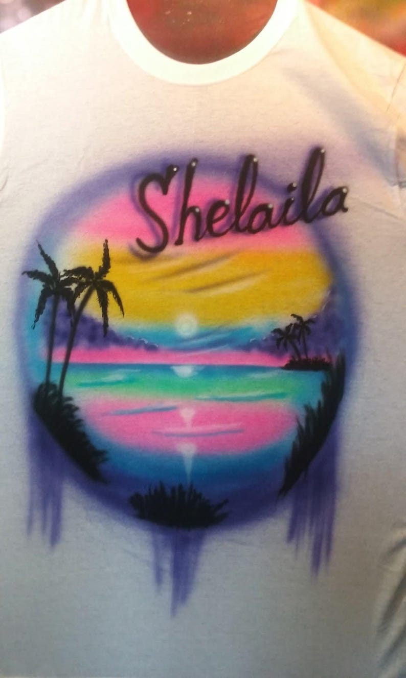 May include: White t-shirt with an airbrushed tropical sunset design. The design features a colorful sky with pink, yellow, and blue hues, palm trees, and the name "Shelaila" in a cursive font.