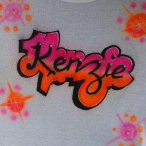 May include: White t-shirt with the name "Kenzie" in a graffiti-style design. The name is in pink and orange with a black outline. The shirt also features pink and orange starburst designs.