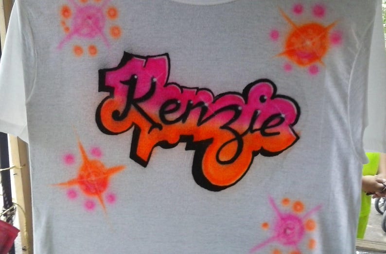 May include: White t-shirt with the name "Kenzie" in a graffiti-style design. The name is in pink and orange with a black outline. The shirt also features pink and orange starburst designs.