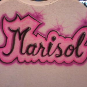 May include: White t-shirt with the name "Marisol" airbrushed in black with a pink outline and accents. The name is stylized with a crown-like design above and a wavy line below, creating a unique, personalized look.