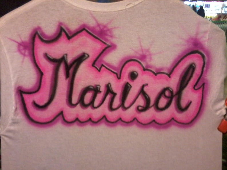 May include: White t-shirt with the name "Marisol" airbrushed in black with a pink outline and accents. The name is stylized with a crown-like design above and a wavy line below, creating a unique, personalized look.