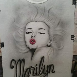May include: White t-shirt featuring a grayscale portrait of Marilyn Monroe with closed eyes and red lipstick. The name "Marilyn" is written in a black, cursive font below the portrait. The shirt is held by black clips.