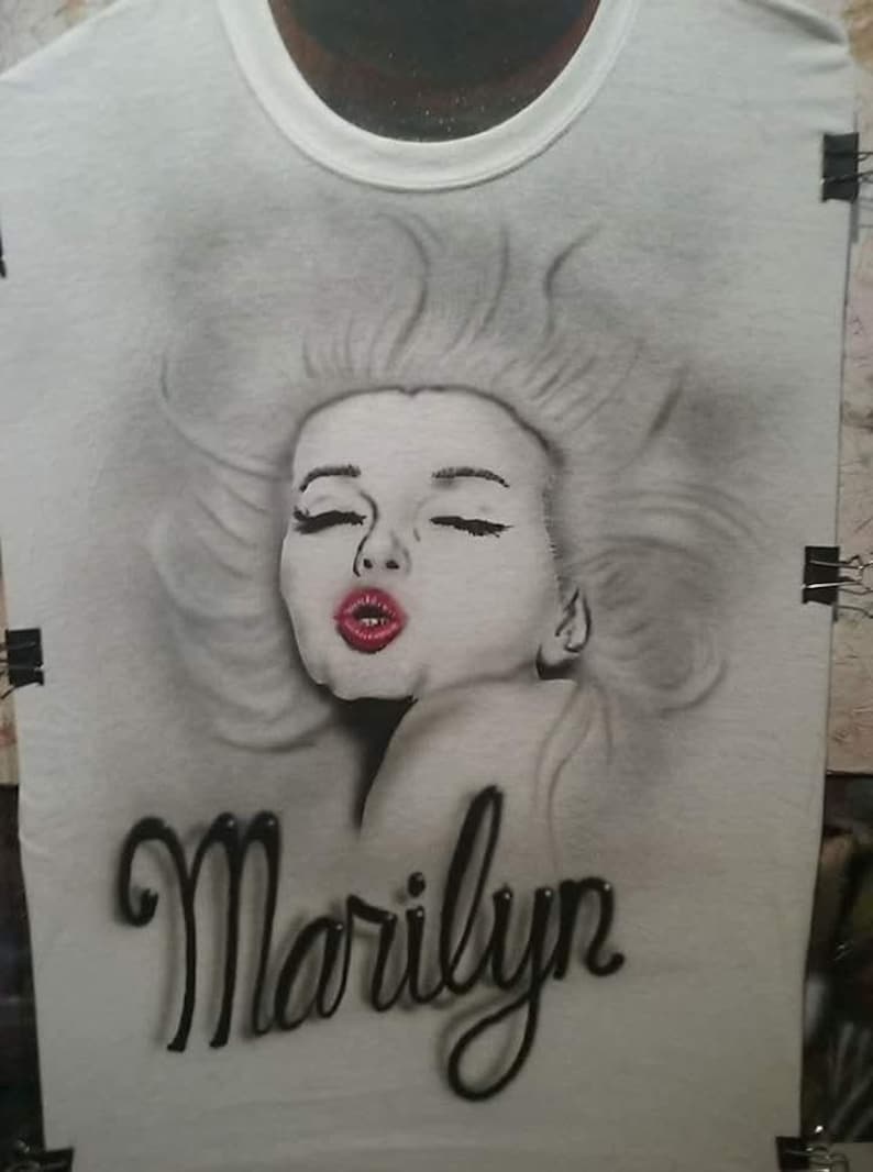 May include: White t-shirt featuring a grayscale portrait of Marilyn Monroe with closed eyes and red lipstick. The name "Marilyn" is written in a black, cursive font below the portrait. The shirt is held by black clips.