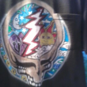 May include: Black t-shirt featuring a colorful Steal Your Face design. The design includes a skull with a lightning bolt, a sun, a moon, and stars. The artwork is in shades of blue, red, yellow, and purple.