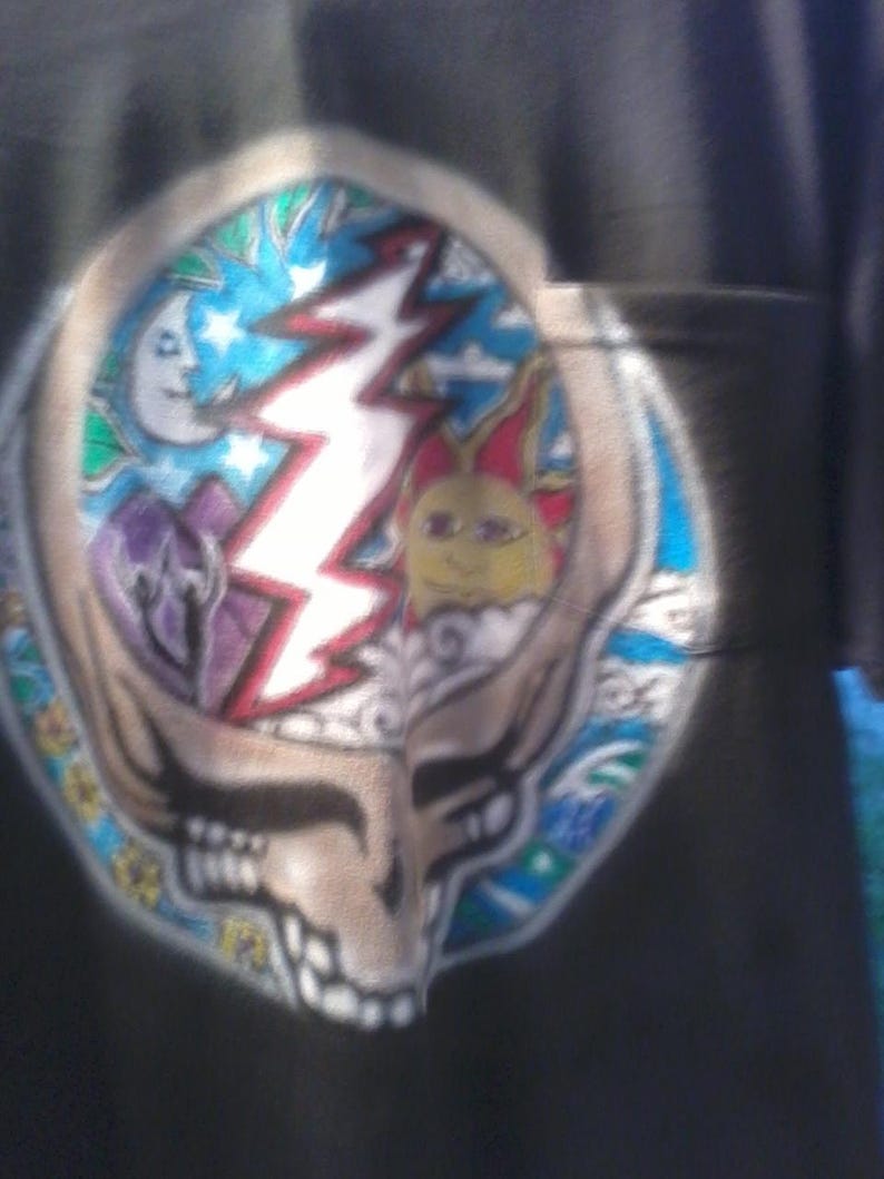 May include: Black t-shirt featuring a colorful Steal Your Face design. The design includes a skull with a lightning bolt, a sun, a moon, and stars. The artwork is in shades of blue, red, yellow, and purple.