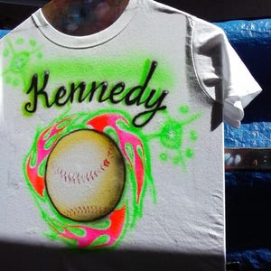 May include: White t-shirt with the name "Kennedy" in black script above a baseball design. The baseball is surrounded by a neon green and pink flame design. The shirt is hanging on a wooden hanger.