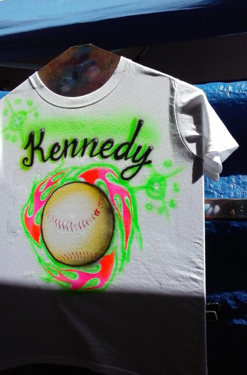 May include: White t-shirt with the name "Kennedy" in black script above a baseball design. The baseball is surrounded by a neon green and pink flame design. The shirt is hanging on a wooden hanger.