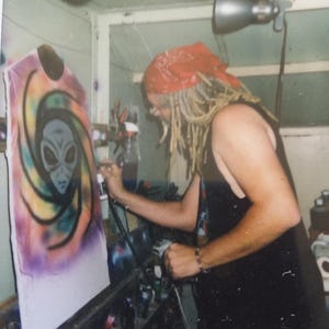 May include: An airbrush artist is shown working on a white t-shirt. The shirt features a colorful design of an alien head within a swirling pattern. The artist is wearing a red bandana and black tank top.
