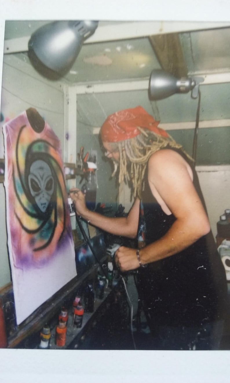 May include: An airbrush artist is shown working on a white t-shirt. The shirt features a colorful design of an alien head within a swirling pattern. The artist is wearing a red bandana and black tank top.
