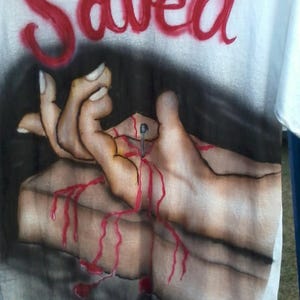 May include: White t-shirt with the word "Saved" in red spray paint across the top. The shirt features a graphic of hands with a nail through them, with red paint dripping down.