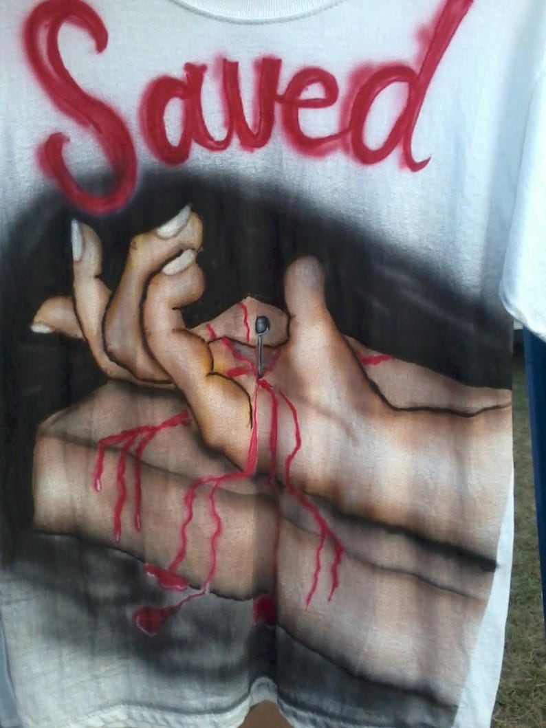 May include: White t-shirt with the word "Saved" in red spray paint across the top. The shirt features a graphic of hands with a nail through them, with red paint dripping down.